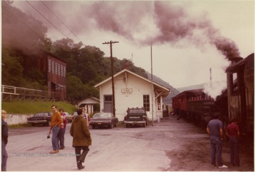 Color photograph of Depot, surrounding buildings and train with steam raised, on the track. Information on the back: "Stephen Trail Collection, Cass, Pocahontas Co. , WV; from Buddy Strokes 3/2000; Photographer Dr. J. W. Strokes, M. D.". 
