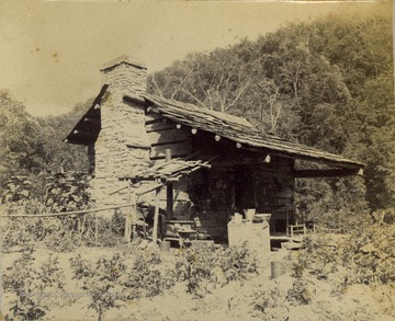 A small split-logged cabin with a stone chimney, surrounded by a vegetable garden.