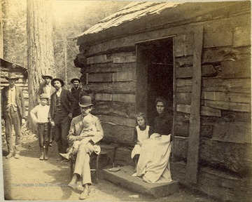 Unidentified woman and little girl sit in the doorway, while a man wearing a bowler hat sits outside holding a baby, with several onlookers behind them. The photo was taken during the construction of the Ohio extension of the Norfolk &amp; Western Railroad.