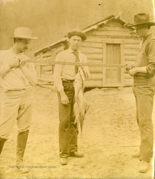 Three unidentified men display a fish catch hanging from a pole. The photograph was taken during the construction of the Ohio extension of the Norfolk &amp; Western Railroad.
