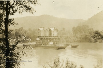 Post card photograph of a large house being transported on a river using scaffolding and pontoon boats. The river is probably the Kanawha or Elk River. 