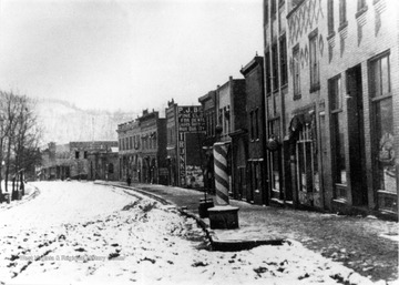 Snow covered Elk Street with a row of shops including a barber's pole in the foreground.