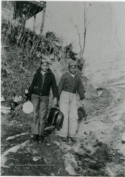 Two African-American boys carrying bag, books and lunch pails. Both are wearing headgear similar to an aviator's leather cap with goggles. Information on p. 121 in "Our Monongalia" by Connie Park Rice. Information with the photograph includes "Courtesy of Kitty Hughes."   