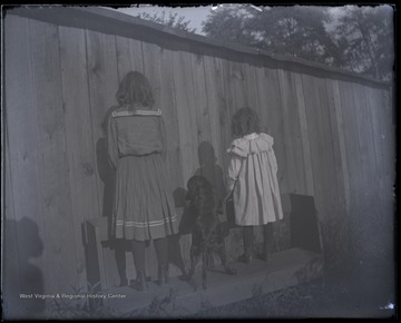 Two unidentified little girls and a dog stand with their backs to the camera.