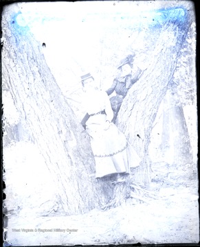 Both unidentified young women wearing early 20th century style lean on a tree for this photograph.