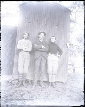 Three unidentified young men pose by an outbuilding. Two are wearing well -padded uniforms, possibly for football.