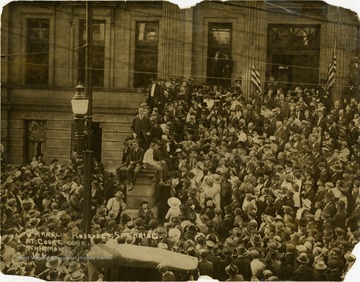 Franklin Roosevelt, standing on a stone column to speak to hundreds of people crowded around the courthouse and in the street. This photograph was most likely taken during the 1920 presidential campaign when Roosevelt was the Democratic Party's vice presidential candidate. Note Roosevelt is standing, without assistance of crutches or braces. He did not contract polio until 1921.