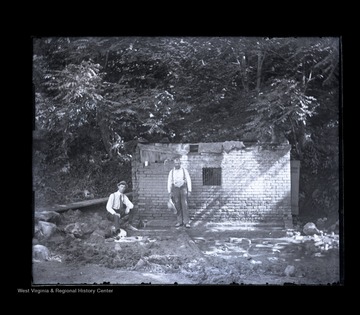 Two unidentified men stand in front of partially fallen structure with bars on its windows -- this may have been a jail.