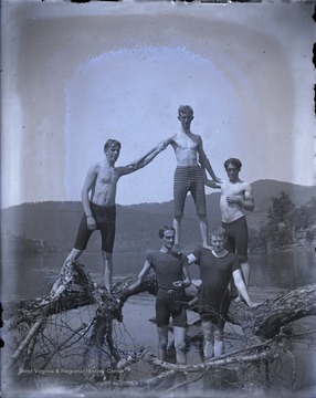 Five unidentified young men wearing swim suits, pose in a pyramid formation, among a fallen tree in a river.