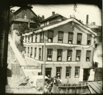 A three story building decorated with bunting and employees (mostly women) standing at several open windows. A delivery wagon, hitched-up and covered with advertisements is park in front.