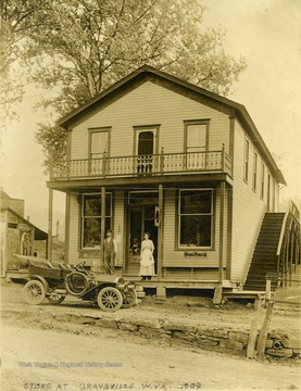 Unidentified man and woman pose in front of the store with a new automobile parked at the curb on a dirt paved road. 