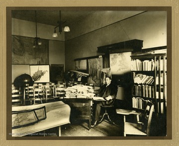 Professor Callahan sits at a desk among maps, books, papers and other other sources of historical documentation.