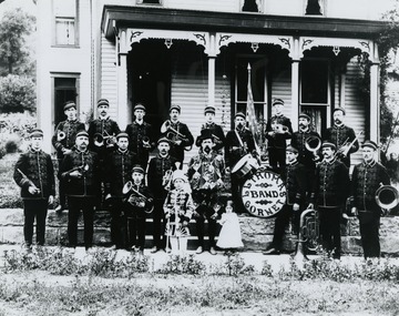 Members identified, back row L to R: ? , Bert Johnson, ? , John Robert Martin, Harry Johnson, Tommy Burke, ___ Collins, Frank Wells, Bill Wells. Front row L to R: ? , Jeff Collins, Bill Morris, Clark Johnson, Lot Thomas, (little girl)Isabelle Thomas, Morris Clovis, Will Howard, Oz Wade. Young boy in front next to Lot Thomas is Ross Collins. Also inscribed on the back : "Lot Thomas furnished the uniforms for the band. Suits were red with gold braids. Mae Johnson Howard made Mr. Thomas' uniform  The men took up a collection and presented it to Lot. Picture taken in front of Lot Thomas home. This home is now the Robert Ownen Funeral Home. At one time this band played for John Phillip Sousa." Labeled on back: "Sara Scott"