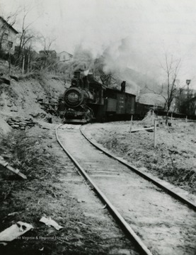 M &amp; W Railway coming around the bend at Scotts Store in Blacksville, West Virginia. Lott Thomas building at left on bank. In distance is Smith Brocks barn and wash house. The smoke hides the Brock home. The bird house at the right is the back of the Thomas property. Photo labeled: Sara Scott