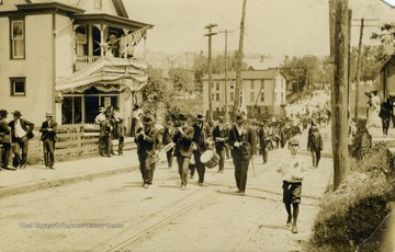Several drummers and men playing the flute lead a parade past eager onlookers watching from the sidewalk and balconies. (From postcard collection legacy system.)