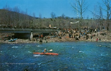 A crowd watches from the river bank as a kayaker begins the 14 mile Championship Race. Published by Gitchell Enterprises. (From postcard collection legacy system.)