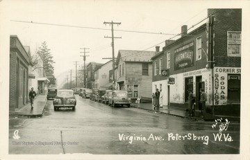 A street lined with cars while pedestrians loiter outside the Corner Service Store. (From postcard collection legacy system.)