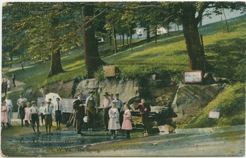 A group of old and young persons stand drinking their beverages around the spring. Published by The Bagley Co. See original for correspondence. (From postcard collection legacy system.)
