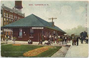 A few men sit by the curb, observing the grounds by the depot as a crowd gathers outside to greet the approaching train. Published by Pike News Company. (From postcard collection legacy system.)