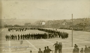 People gather on the field while a small band plays in the middle during halftime of a game. See original for correspondence. (From postcard collection legacy system.)