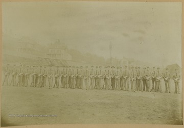 Group portrait of unidentified WVU cadets lined up on the athletic field. Houses on High street can be seen in the background. 