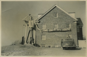 House displays banner reading, "Come on-a my house" while a giant crafted man the size of the house stands awaiting guests.