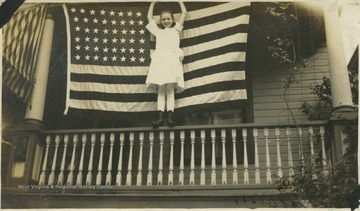 Young girl stands on the porch railing and stretches in front of the large American flag.
