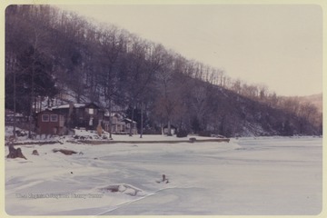 View from the snow covered, frozen river showing a house beneath the woods.
