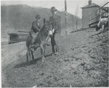 Unidentified men pose with a mule. A train car can be seen in the background. 