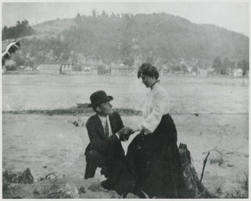 A man holds a woman's hand on the bank of New River. Avis can be seen in the background.