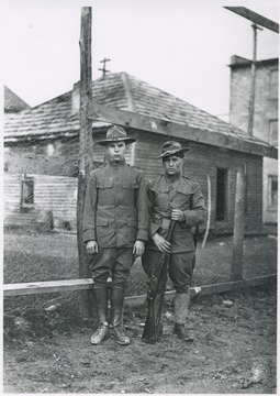 Two men in uniform pictured next to a fence. One carries a rifle. Subjects unidentified. 
