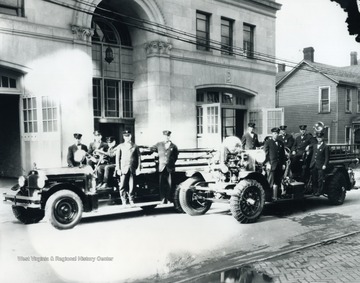 Print number 1265. Trucks are in front of the fire department building on Spruce Street. Doc, the firehouse dog, blends in among the Firemen standing on right vehicle.
