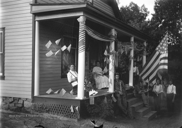 Woman on left is Grace Green (sister to Green brothers), man on porch furthest back is Thomas Benton Green (homeowner), woman next to him is Mary Rupert Green, boy holding American flag on far right is Ray Green, and young man in foreground leaning against post is John Green. The rest of the people are neighbors on hill above the town.