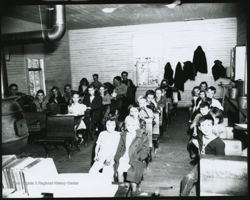 Looking from the front of the classroom, school children of various ages, are pictured sitting at their desks. Subjects unidentified. 