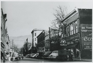 Parked cars line the street in front of the store buildings. A. W. Cox Department Store pictured on the right. 