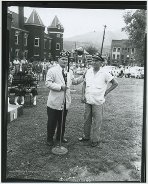 Two men stand by a microphone in the yard. Musicians are pictured with instruments seated behind music stands on the left. A small crowd can be seen in the background watching the event. Subjects unidentified. 