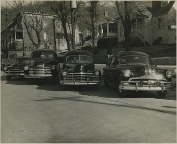 Cars parked along the street in front of residences. The automobile in the center sports a sign reading "Ambulance" on its dashboard. To the far right, a sign for Barnett Funeral Home is pictured partially.