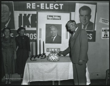 An unidentified man cuts a cake to celebrate the birthday of President Dwight D. "Ike" Eisenhower. In the background hangs a campaign poster to re-elect Eisenhower as president with Richard Nixon as his vice-president. 