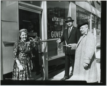 An unidentified man gets ready to cut the ribbon blocking the entranced to the building. On the window reads, "Democratic Headquarters for Wally Barron for Governor". The man in the center with his hand held out is Richard Baylor. The woman to the left is unidentified. 