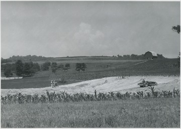 A group of unidentified men watch on the left as a tractor digs up the land, perhaps for a reservoir.
