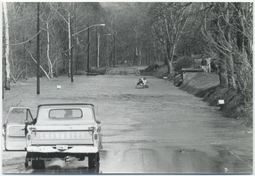 A Chevy truck is stopped in front of a submerged part of a road while two unidentified persons row toward it. 