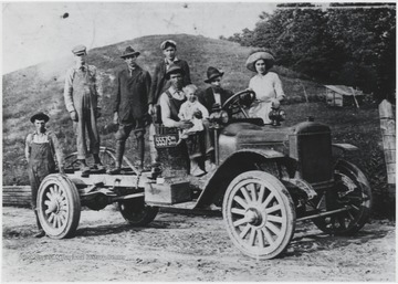 A group of men, dressed in overalls and working boots, stand on the back of the automobile. A man holding a child sits in the passenger seat with an associate driving. A woman is also pictured behind the car. Subjects unidentified. 