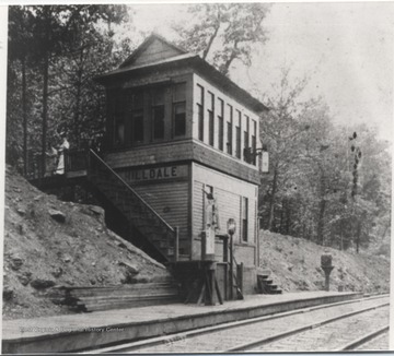 Looking at the station building from across the train tracks. An unidentified woman is pictured looking over the staircase railing. 