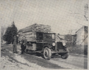 Two unidentified men stand beside a truck loaded with lumber. 