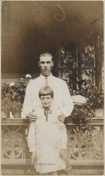 A man with his hair slicked back stands behind a young girl in a dress, gripping her shoulders. The two unidentified persons stand in front of floral pots arranged outside a house's balcony.