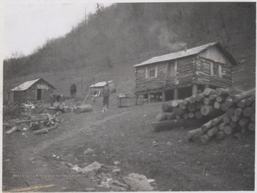 Two unidentified men are pictured outside the log home. One chops wood while the other stands beside a dog. 
