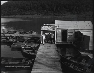 Three men stand beneath a sign that reads, "Welcome--Bluestone Boat Dock No. 1". Empty row boats surround the dock. The man standing at the right is Gordon Skaggs, the dock owner.