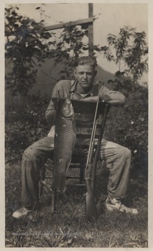 Foy sits on a chair while showing off a large fish and rifle. The boy is son to W. M. Meador. His brother, the photographer, is Luther Meador. 