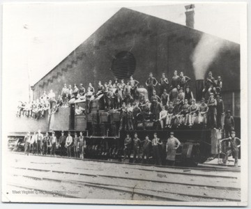 Engine No. 7 sitting beside stall No. 1 of the roundhouse. A group of unidentified workers stand on along the tracks and sit on the train. 
