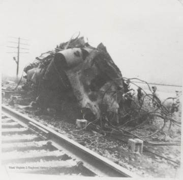 Looking at the toppled engine from the railroad tracks. To the right, a group of unidentified workers observe the damage. 